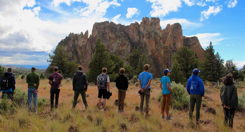 A group of people stand in a line, facing away from the camera. They appear to be looking at a rock formation in the distance.